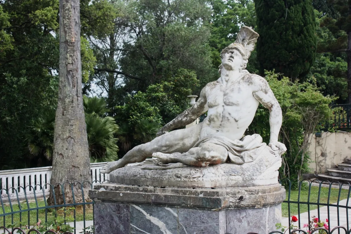 A white marble statue of a wounded warrior in a crested helmet, leaning back on a stone base among garden trees (the Dying Achilles at the Achilleion palace on Corfu, not the show)