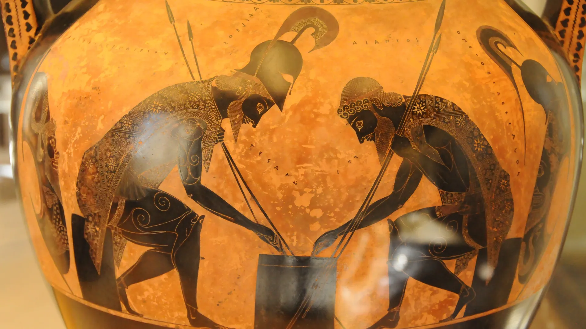 Two armoured warriors crouch over a game on an orange Greek vase, painted in black figures with their spears crossed between them