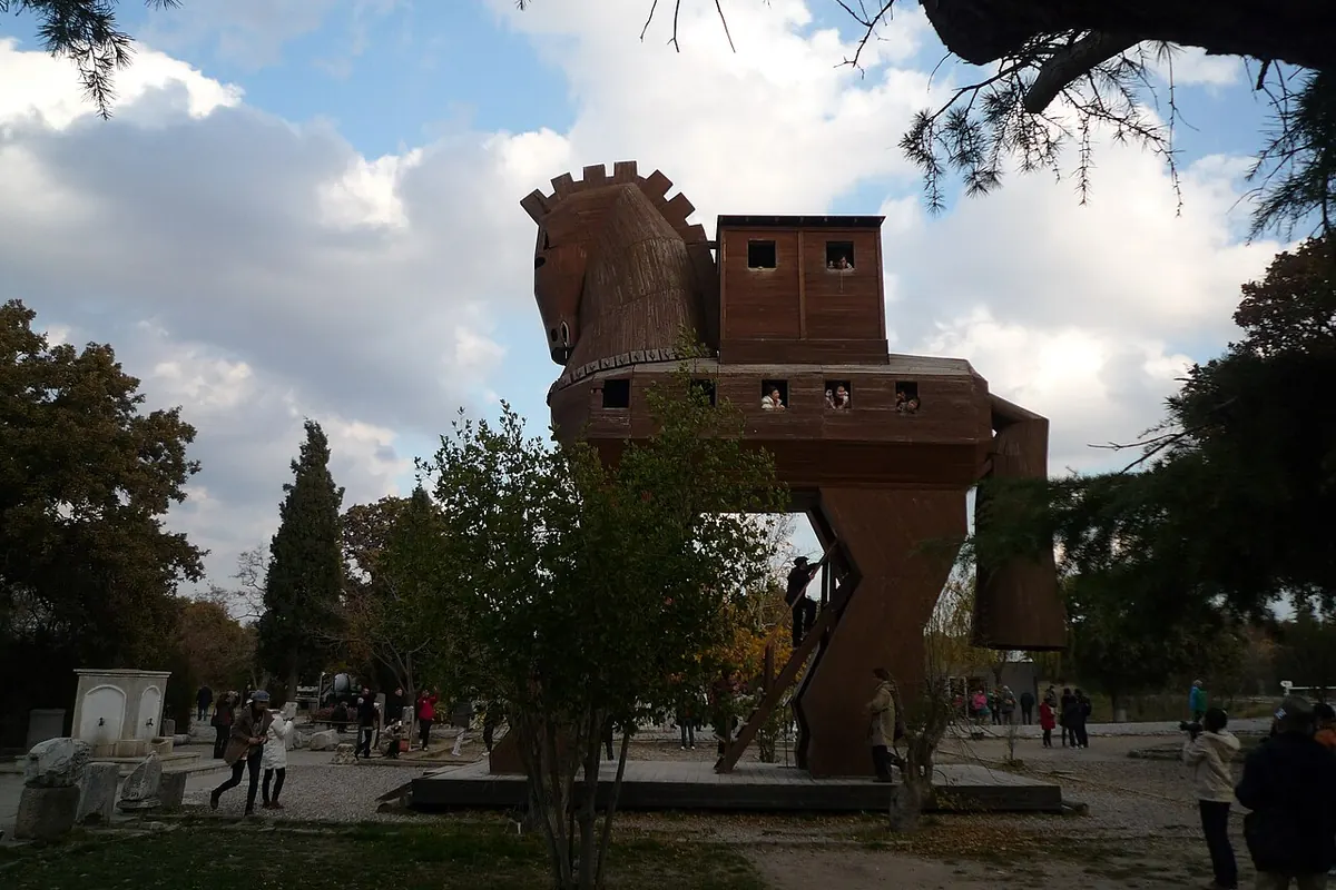 A tall wooden horse with a castle-like platform on its back stands among pine trees under a cloudy sky, with visitors walking below (the Trojan horse replica at the archaeological site of Troy in Turkey, not part of the show)